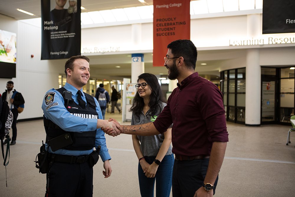 A police officer from the auxiliary unit shakes hands with a man at Fleming university in front a woman who is standing nearby